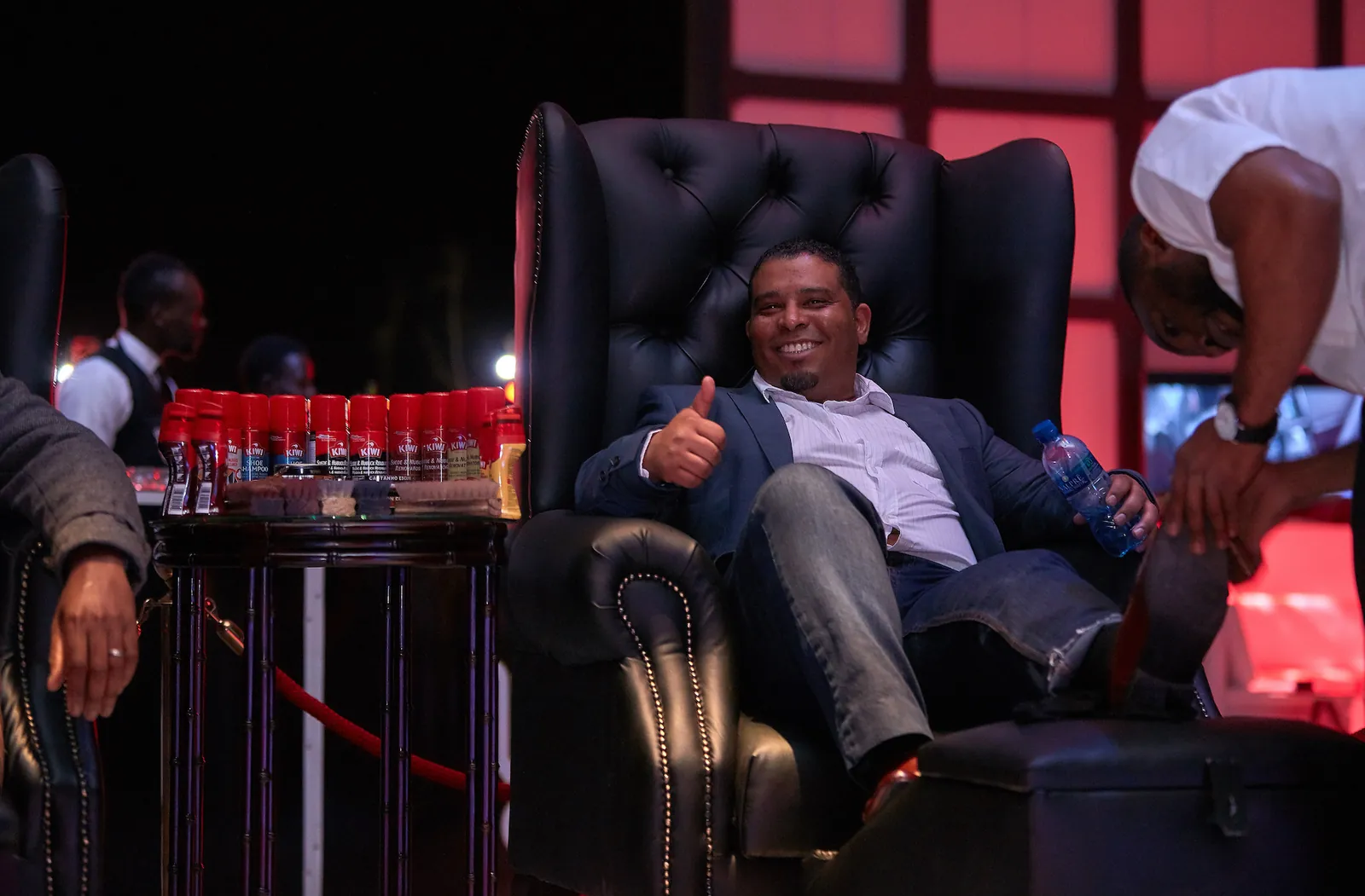 A smiling guest gives a thumbs up from a leather wingback chair while a Madinda Shoe Shine specialist polishes his shoes, beside a table of Kiwi products.