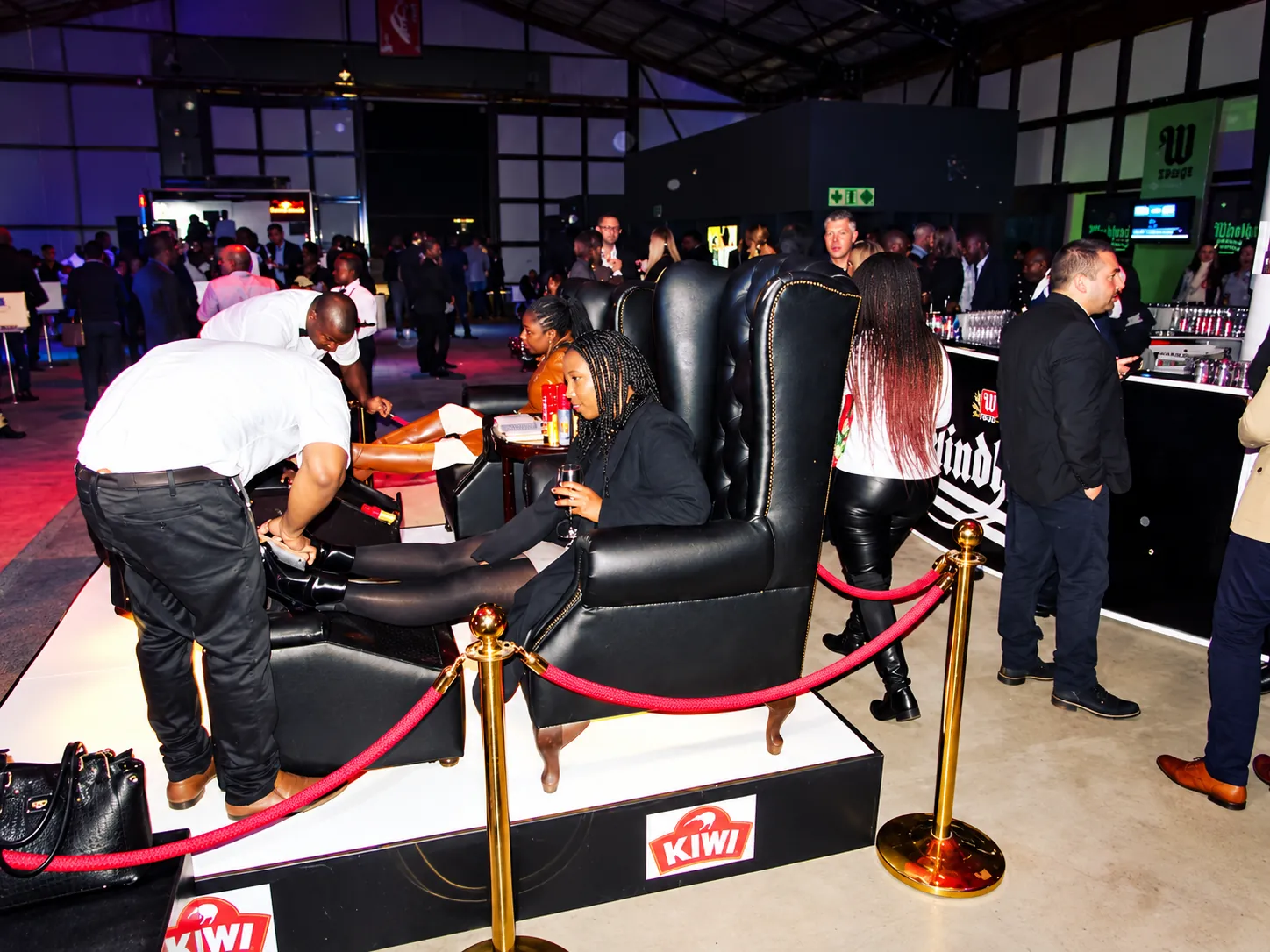 Madinda Shoe Shine specialists polish guests’ boots in black wingback chairs on a roped-off platform at an evening corporate event.